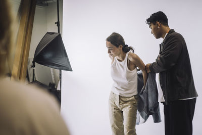 Male designer helping fashion model wearing jacket during photo shoot in studio