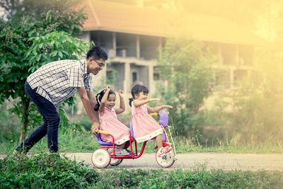 Full length of sisters riding tricycle with father on road at public park