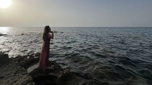 Rear view of woman standing at beach against clear sky during sunset