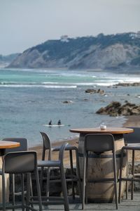 Empty chairs and table at beach against sky