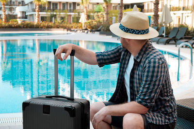 Caucasian man wearing sunhat with suitcase, baggage coming to hotel, looking at empty swimming pool