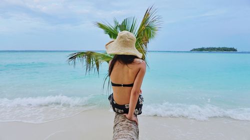Rear view of woman sitting on coconut palm tree at beach against sky