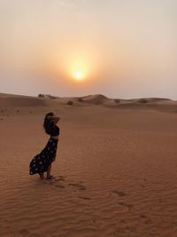 Side view of woman standing at desert against sky during sunset