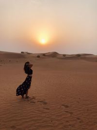 Side view of woman standing at desert against sky during sunset