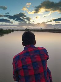 Rear view of man standing by lake against sky during sunset