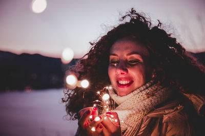 Smiling woman holding illuminated lights against sky