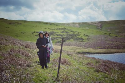 Full length of woman with children walking on grassy field at hill against cloudy sky