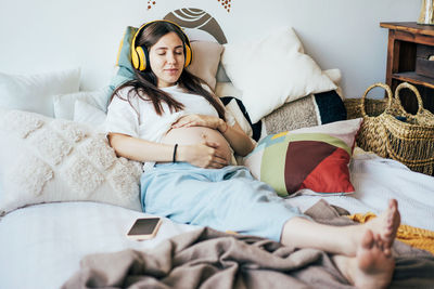 Young happy pregnant modern woman resting in pleasure on the bed listening to music on headphones. 