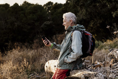 Senior man using mobile phone on wooden log in field during sunset