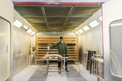 Male carpenter with protective face mask spraying paint on wood material in spraybooth