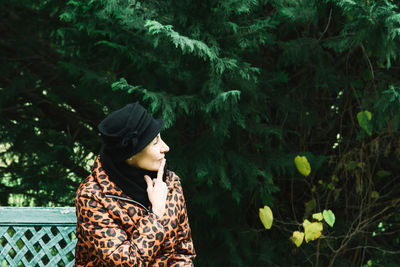 Woman looking away while sitting on bench against trees