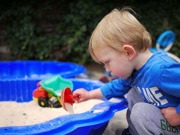 Side view of cute boy playing with sand in blue container at back yard