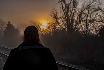 Rear view of woman standing by bare trees during sunset