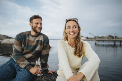 Happy woman sitting with male friend having wine while sitting on rock near lake