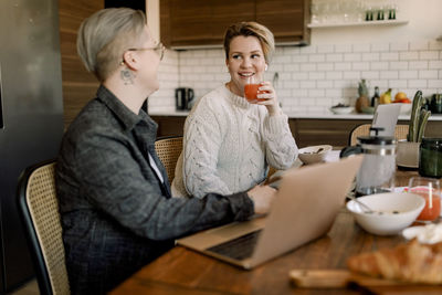 Young lesbian woman drinking juice while looking at girlfriend sitting at dining table in kitchen