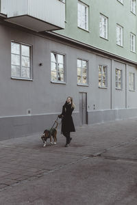 Full length of young woman walking with dog on footpath by building in city