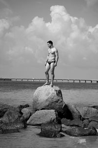 Full length of shirtless man in underwear standing on rock amidst sea against sky