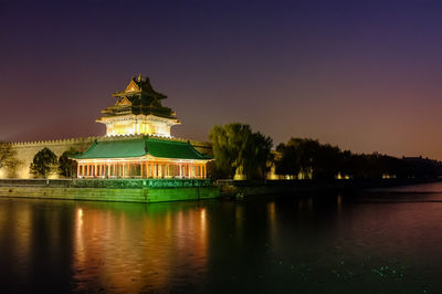 Illuminated building by lake against sky during sunset