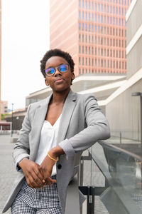 Confident african american female entrepreneur in eyeglasses and formal jacket standing on glass fence in street