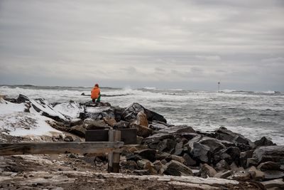 Rear view of man crouching on rock formation at beach against sky