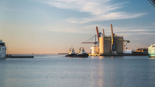 Sunset seaport scene with cargo ships, tugboats, cranes, and serene reflective water 