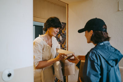 Mature woman doing digital signature while receiving parcel from delivery person at house