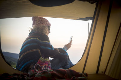 Smiling woman using smart phone while sitting in tent against sky
