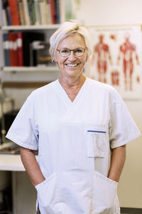 Portrait of happy senior orthopedic surgeon standing with hands in pockets in clinic