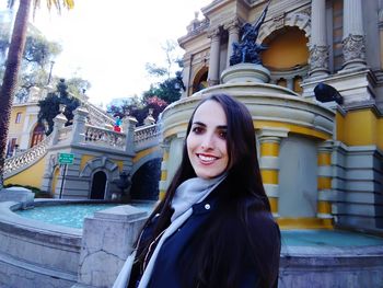 Portrait of smiling young woman against fountain and building