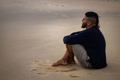 Side view of young man sitting on sand at sandy beach