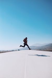 Full length of man jumping over road against clear blue sky on sunny day