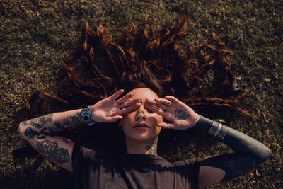 From above serene stylish brunette in casual clothes with tattoos and piercing laying on grassy glade and covering face with hands while relaxing alone in park