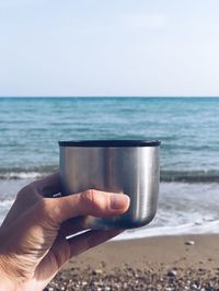 Close-up of hand holding drink in container at beach against clear sky