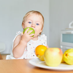 Blond blue-eyed toddler biting green aple and looking at camera. plate full of fruits