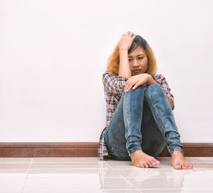 Full length of depressed young woman sitting on tiled floor against white wall