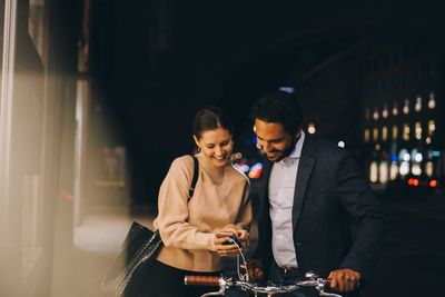 Smiling woman showing smart phone to male friend with bicycle while standing in city at night