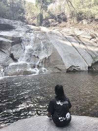 Rear view of woman sitting on rock against waterfall