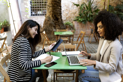 Businesswoman writing in book while female coworker using laptop sitting at cafe