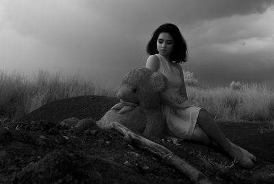 Young woman with teddy bear sitting on field against sky