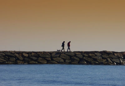 Mid distance view of couple with dog walking at rocky beach against sky during sunset