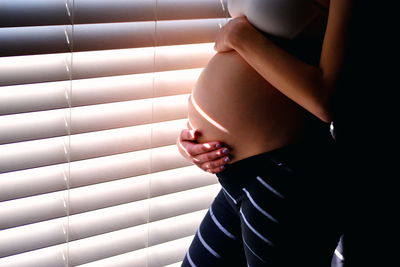 Midsection of pregnant woman standing by blinds at home