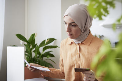 Smiling woman with hijab holding credit card while doing online shopping