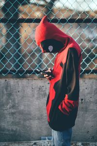 Side view of man using phone while standing against fence