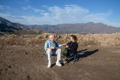 Woman and her grandfather talking while sitting on camping chairs outdoors in nature.