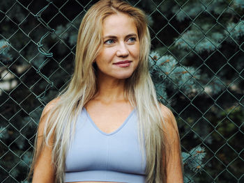 Young beautiful blond woman with long hair in light blue activewear smiling with wire mesh on backgr