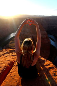 Rear view of young woman making heart shape while sitting on rock