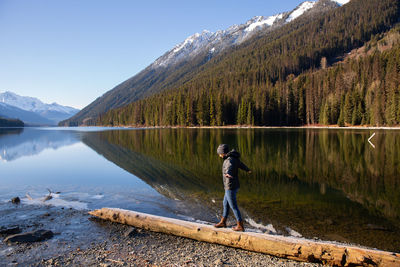 Side view of young woman walking on log at lakeshore against mountain