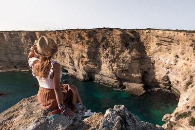 Side view of woman sitting on rock against sky