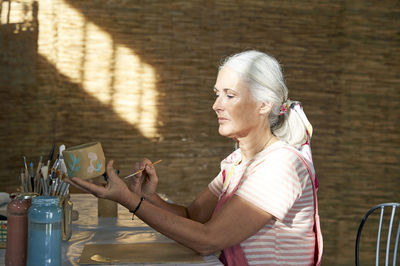 Woman painting clay pot sitting on chair at table
