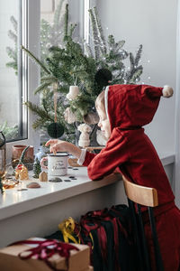 A thoughtful boy in a santa costume sits at the window and makes christmas decorations